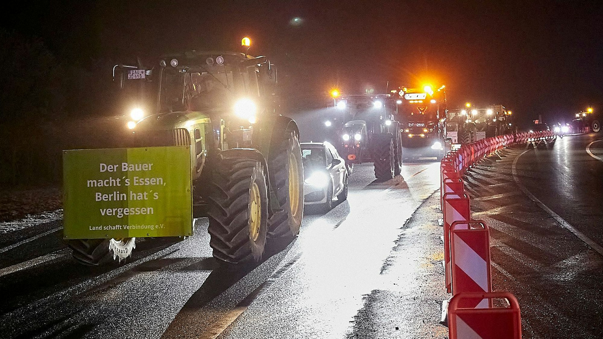 Ein Traktor mit einem Protestplakat fährt im Dunkeln über eine Straße. Hinter ihm sind zahlreiche weitere Fahrzeuge auf dem Weg zum Mahnfeuer in Blankenheim.