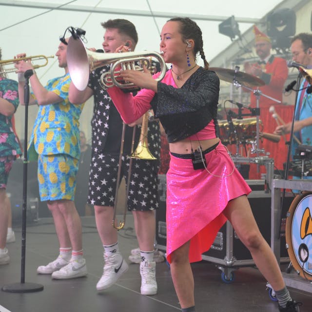 Die Musikerinnen und Musik der Band Druckluft stehen in ihrer bunten Kleidung auf der Bühne, unter anderem mit Posaunen und Trompeten.