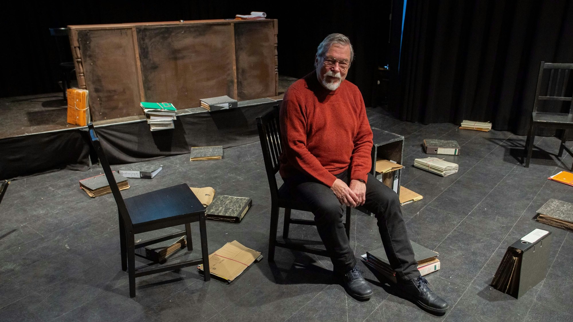 Raimund Binder sitzt auf einem Stuhl auf einer Bühne. Um ihn herum im Bühnenbild herrscht Chaos. Überall liegen Kladden und Ordner.