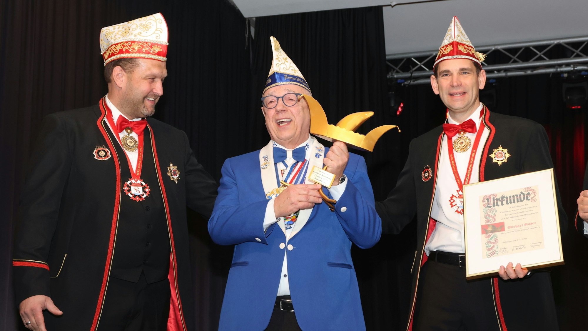 Drei Karnevalisten stehen beisammen. Der Mann in der Mitte hält eine Auszeichnung in Form einer goldenen Narrenkappe in den Händen.