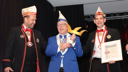 Drei Karnevalisten stehen beisammen. Der Mann in der Mitte hält eine Auszeichnung in Form einer goldenen Narrenkappe in den Händen.