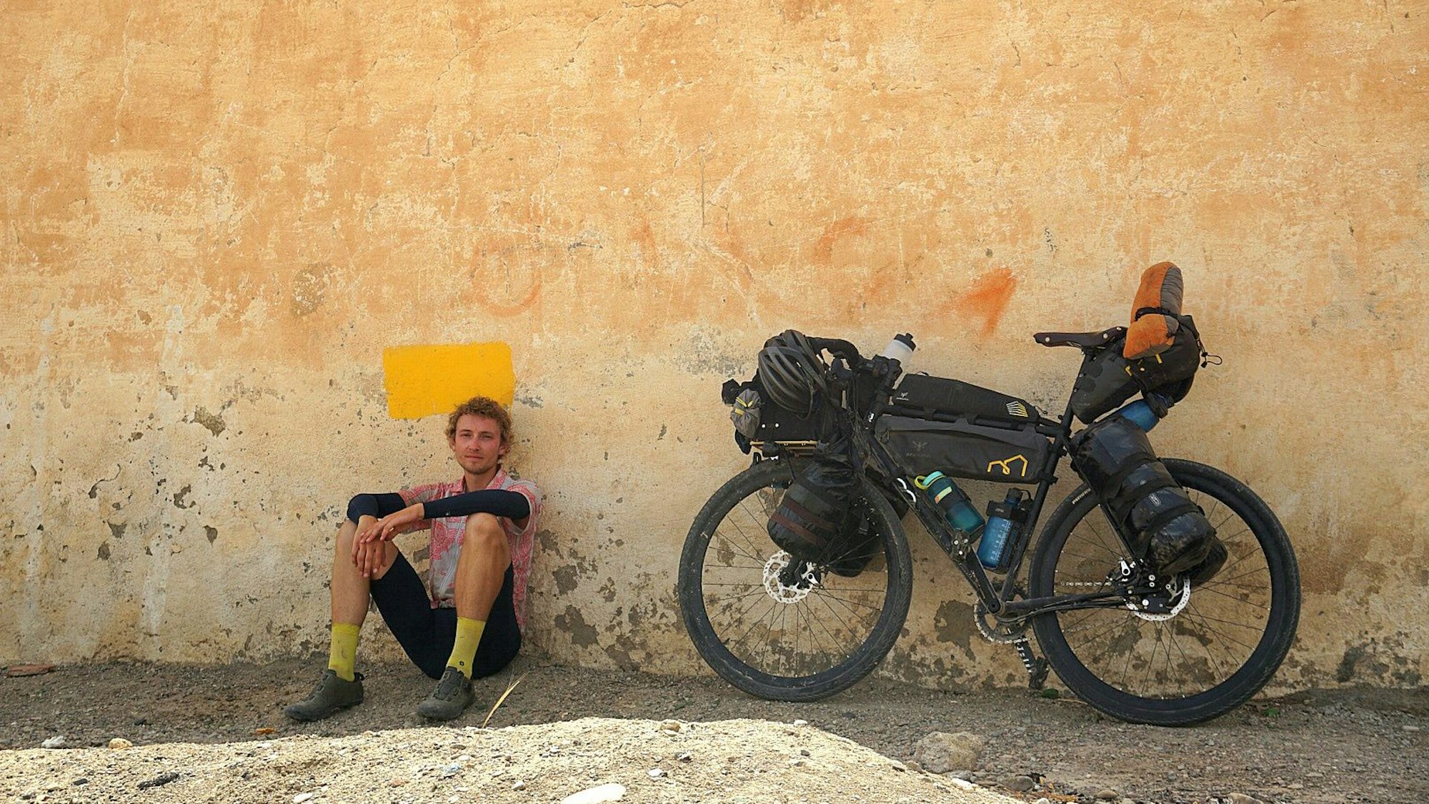 Ein Mann sitzt vor einer Mauer im Schatten, daneben steht sein Fahrrad.