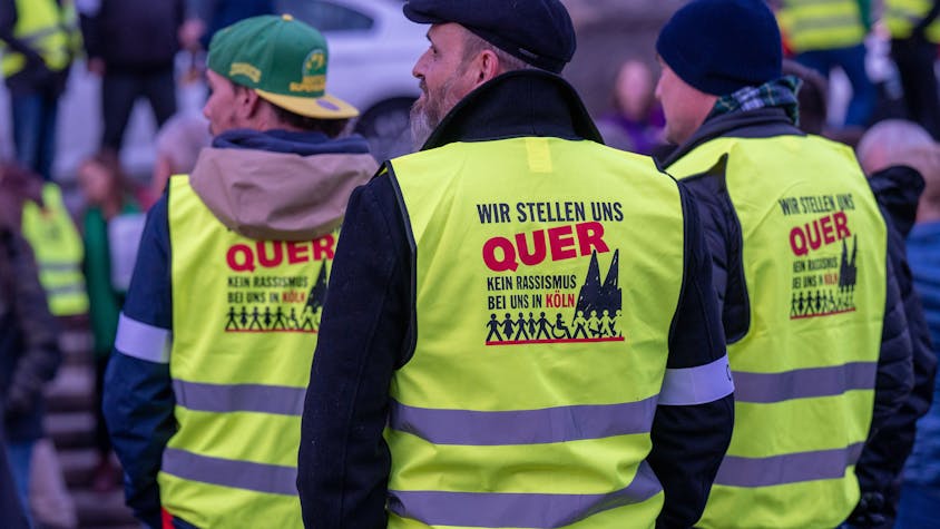 Drei Männer in gelben Warnwesten mit der Aufschrift „Wir stellen uns quer. Kein Rassismus bei uns in Köln“