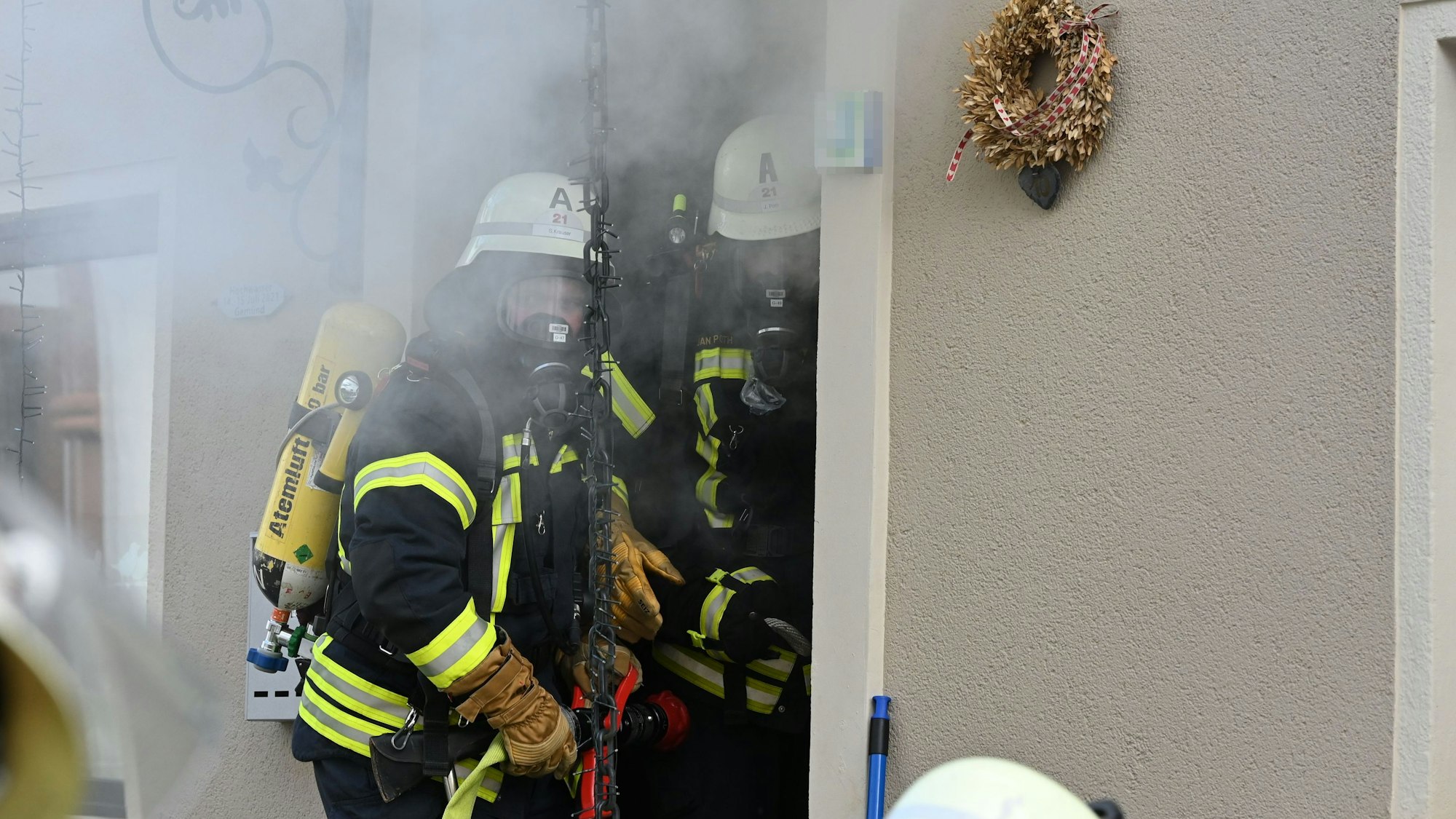 Zwei Feuerwehrleute tragen Atemschutzgeräte. Sie machen sich bereit, in ein Haus zu gehen, aus dem Rauch quillt.