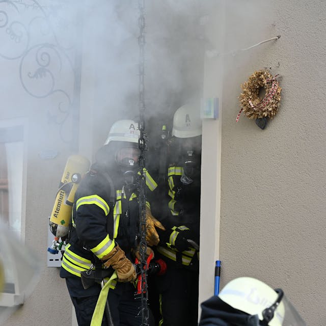 Zwei Feuerwehrleute tragen Atemschutzgeräte. Sie machen sich bereit, in ein Haus zu gehen, aus dem Rauch quillt.&nbsp;
