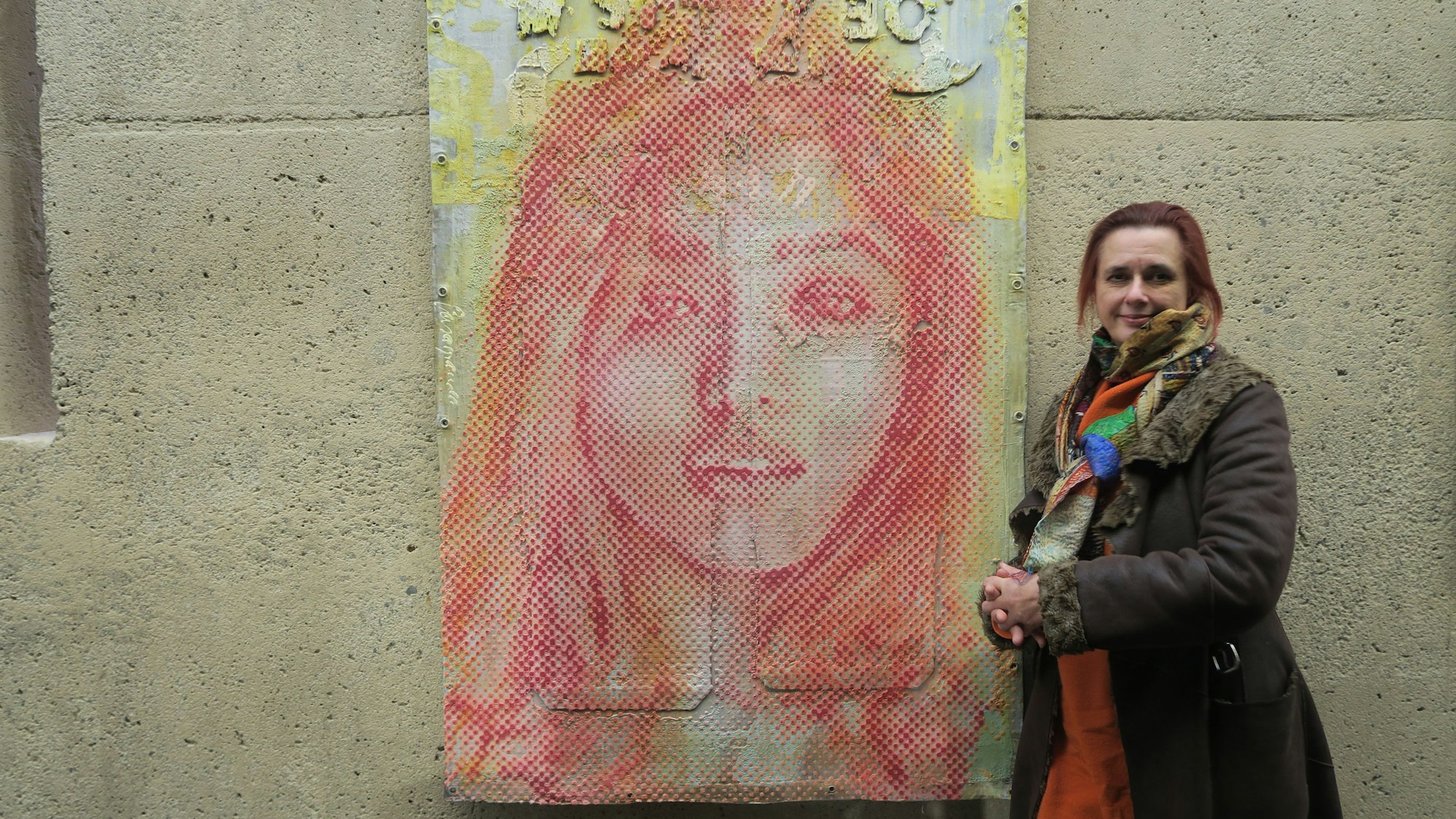 Eine Frau im braunen Mantel lehnt an einer Wand neben einem großformatigen Porträt eines Mädchens.