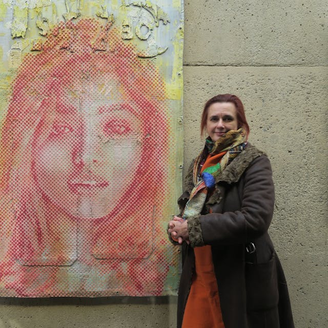 Eine Frau im braunen Mantel lehnt an einer Wand neben einem großformatigen Porträt eines Mädchens.