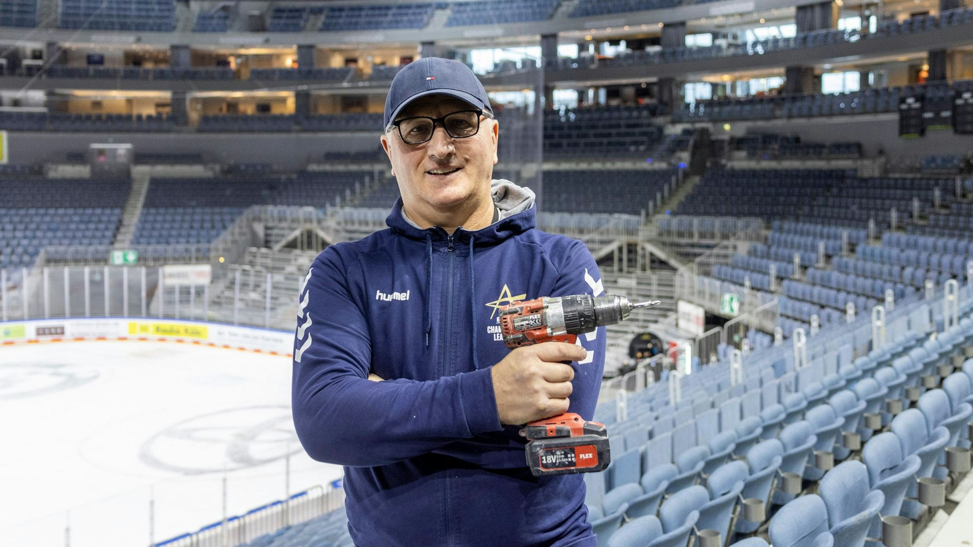 Hausmeister Uzeir Hadzovic steht mit einem Akkuschrauber in der Lanxess-Arena. Im Hintergrund ist noch die Eisfläche zusehen.
