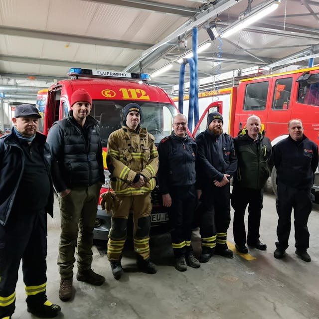 Das Foto zeigt eine Gruppe von Feuerwehrleuten in der Fahrzeughalle der Freiwilligen Feuerwehr Mechernich vor zwei Einsatzfahrzeugen.