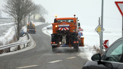 Ein Räumfahrzeug des Winterdienstes befreit eine Straße in der Eifel von Schneemassen. Weitere Schneeflocken fallen vom Himmel. (Symbolbild)