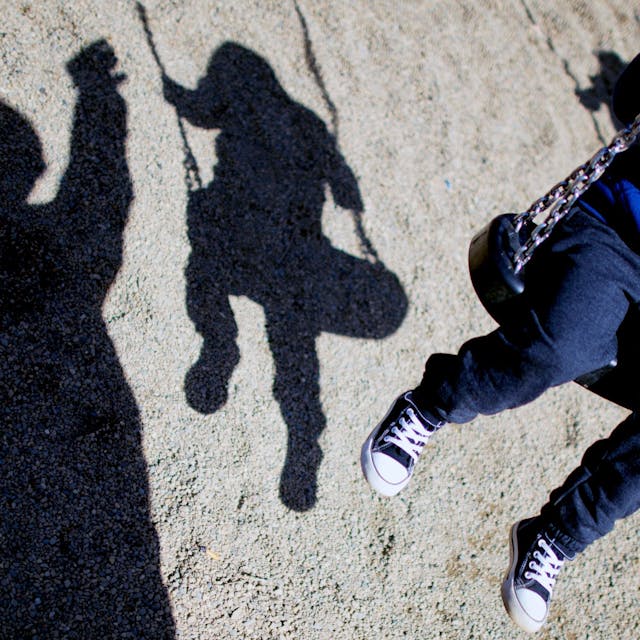 Ein Kind sitzt auf einer Schaukel, sein Schatten ist auf dem Boden zu sehen, ebenso der Schatten einer weiteren Person.