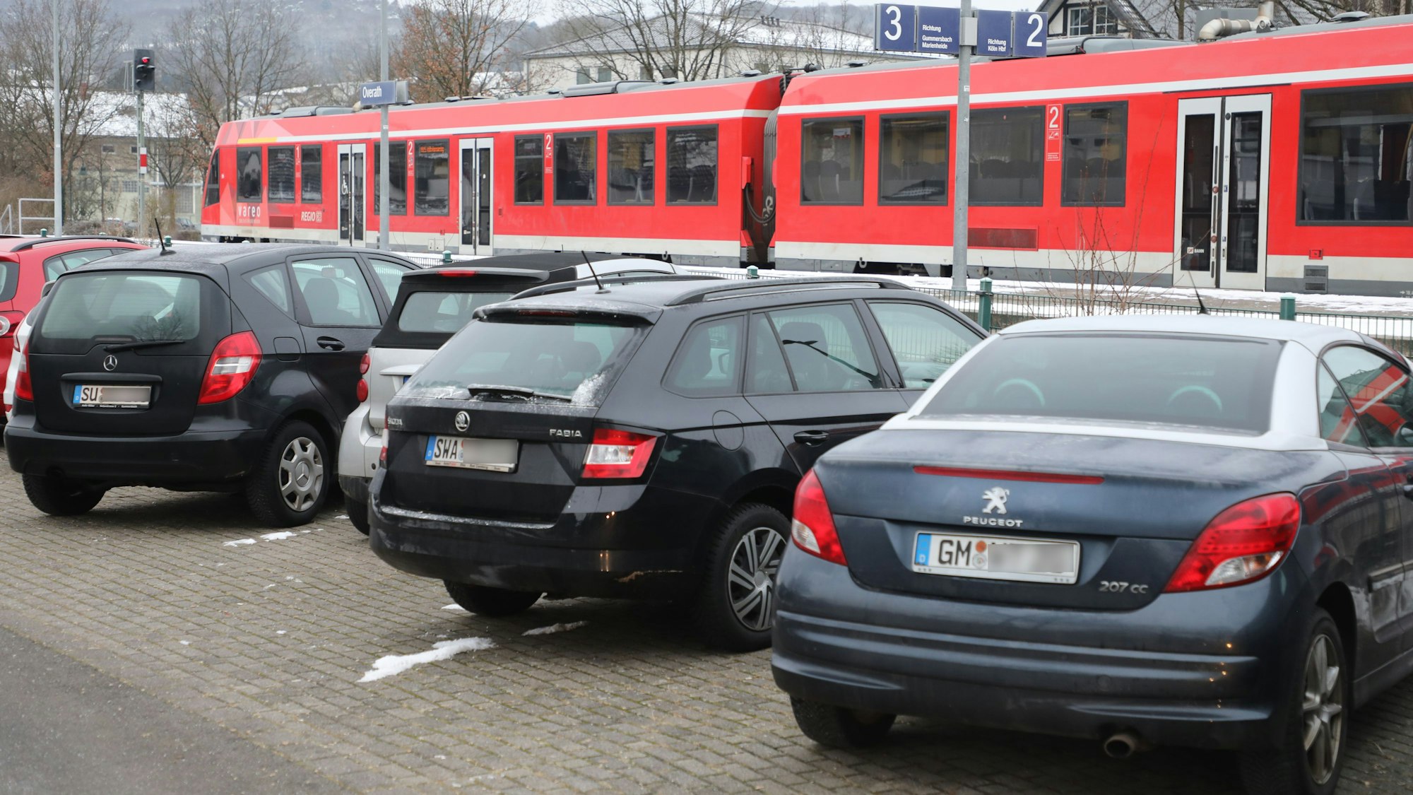 Autos stehen auf einem Parkplatz am Overather Bahnhof, dahinter ist eine abfahrbereite Regionalbahn zu sehen.