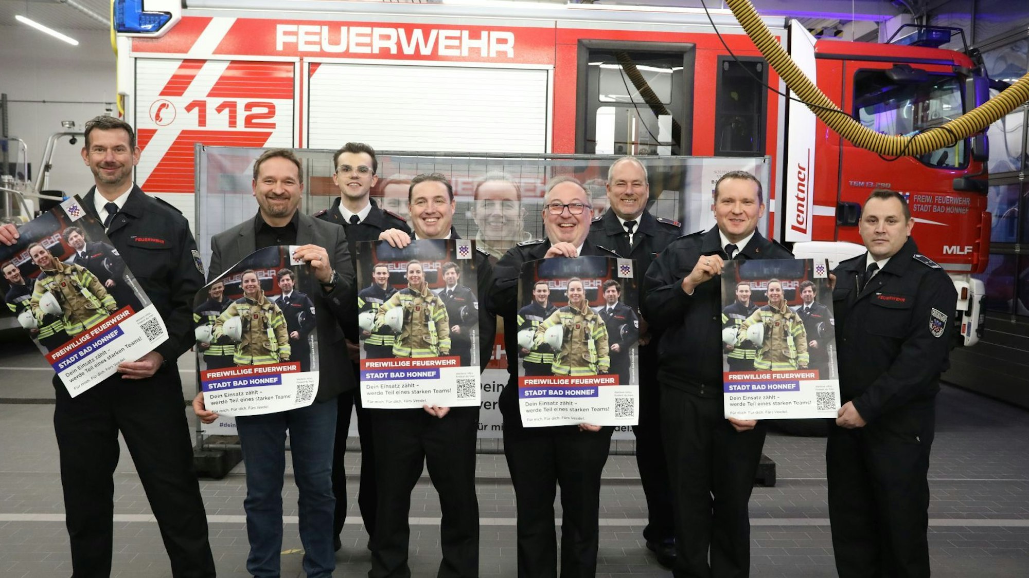 Eine Gruppe von acht Männern steht vor einem Feuerwehrfahrzeug. Sie halten Plakate in die Kamera.
