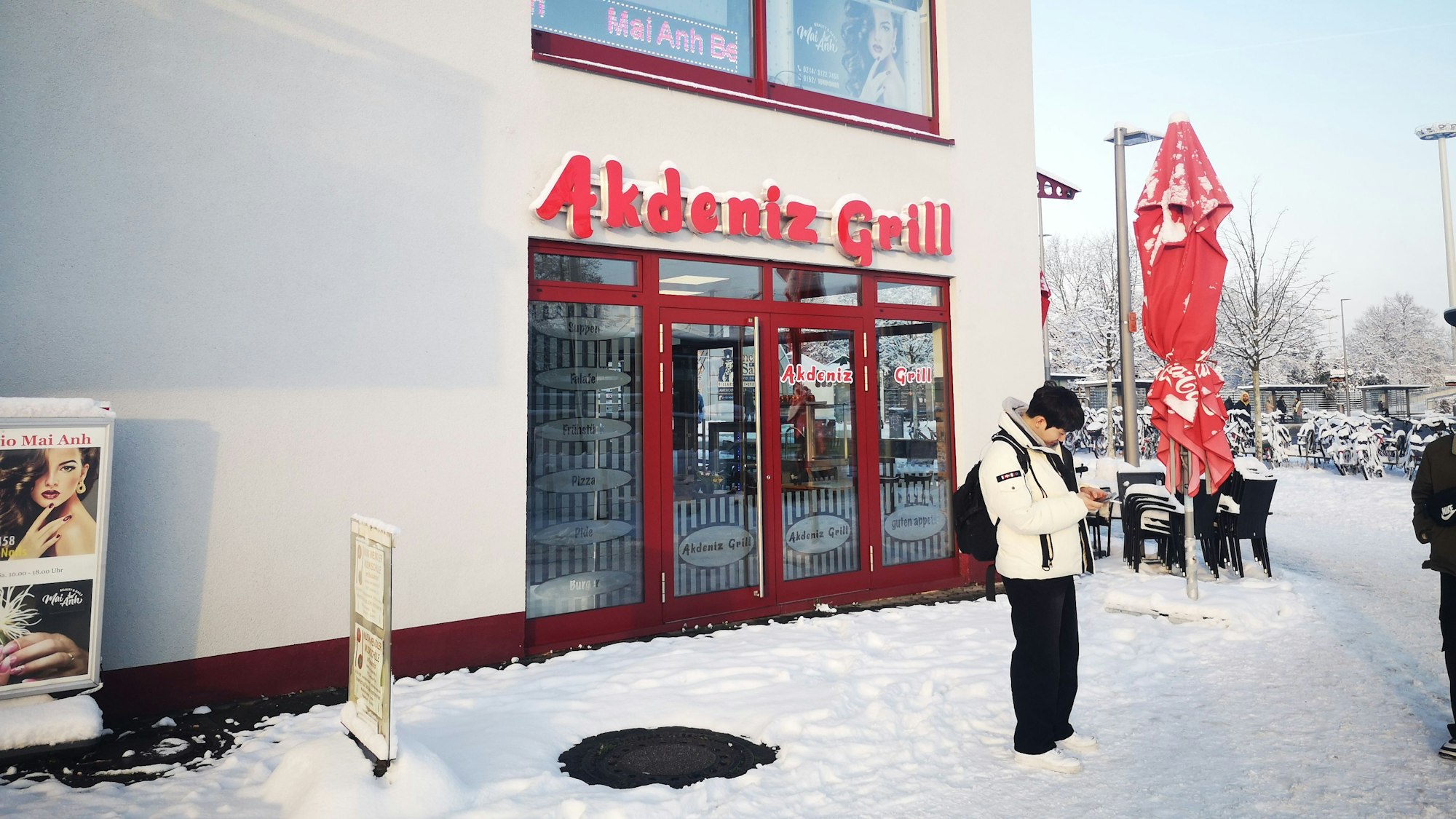 Der Akdeniz Grill in Leverkusen Mitte in der Außenansicht. Es liegt Schnee. Vor dem Restaurant schaut ein junger Mann auf sein Handy. Links unten im Bild befindet sich ein Werbeschild, oben rechts ein ausziehbarer Schirm.