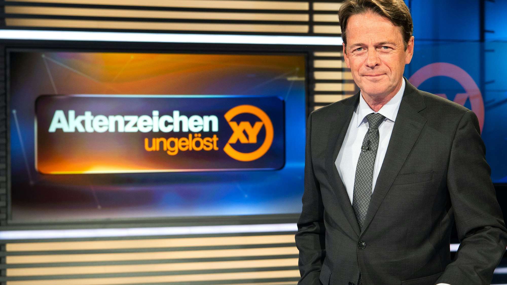 ZDF-Moderator Rudi Cerne steht im Studio vor dem Logo der Sendung „Aktenzeichen XY ... ungelöst“.