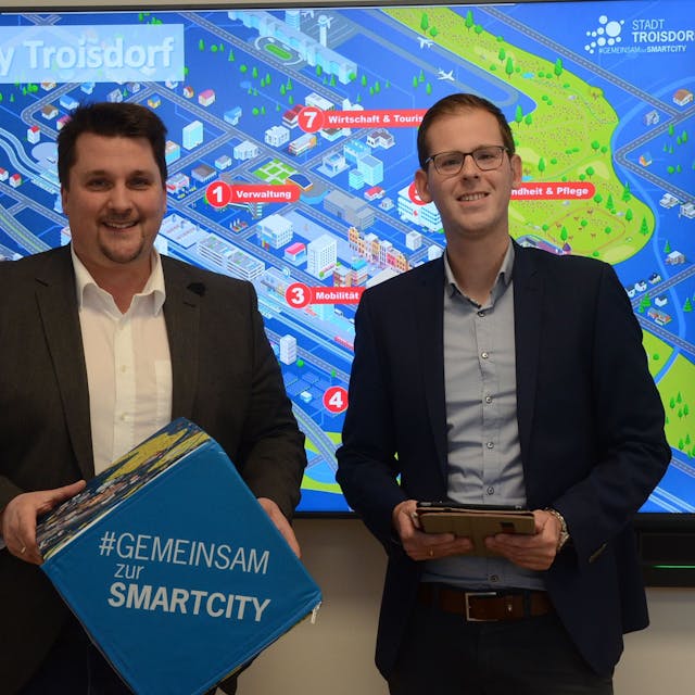 Zwei Männer stehen vor einem bunten Schaubild mit der Überschrift Smart City Troisdorf. Einer der Männer hält einen blauen Würfel, der andere einen Tablet-Computer