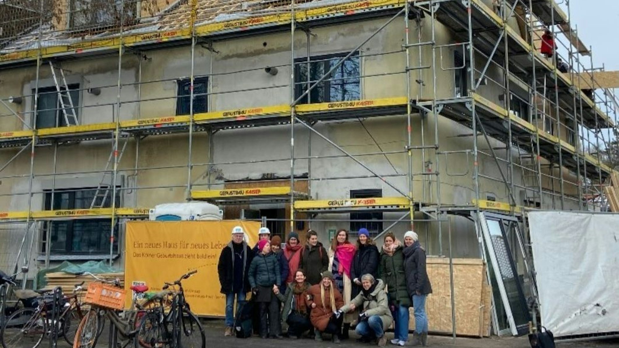 Eine Gruppe Menschen mit und ohne Baustellenhelm steht vor einem eingerüsteten Eckhaus.