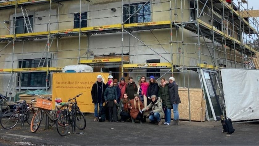 Eine Gruppe Menschen mit und ohne Baustellenhelm steht vor einem eingerüsteten Eckhaus.
