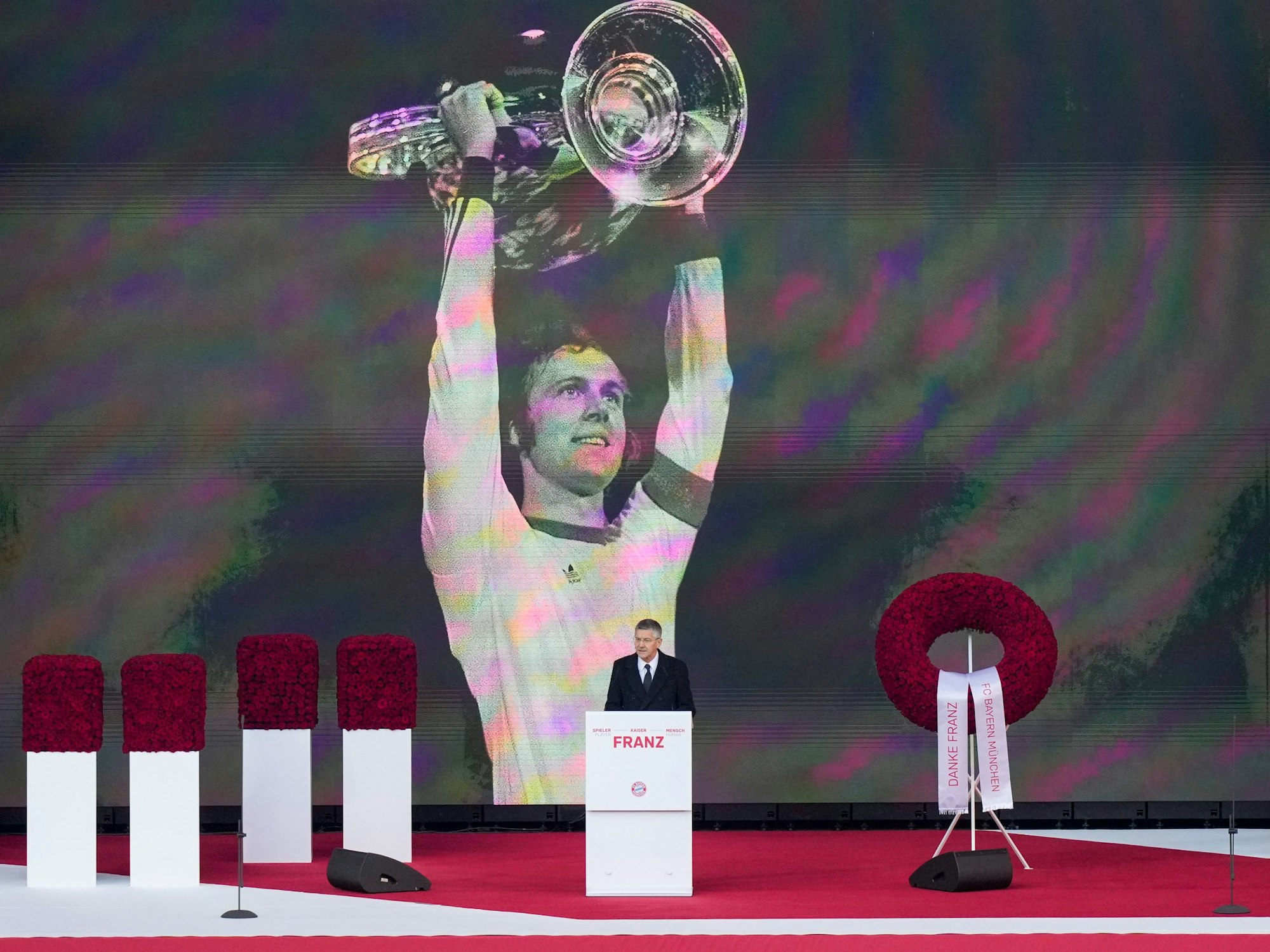 Bayern Münchens Vereinspräsident Herbert Hainer hält bei der Gedenkfeier für Franz Beckenbauer eine Rede. Im Hintergrund ist ein überlebensgroßes Bild von Beckenbauer mit Pokal.
