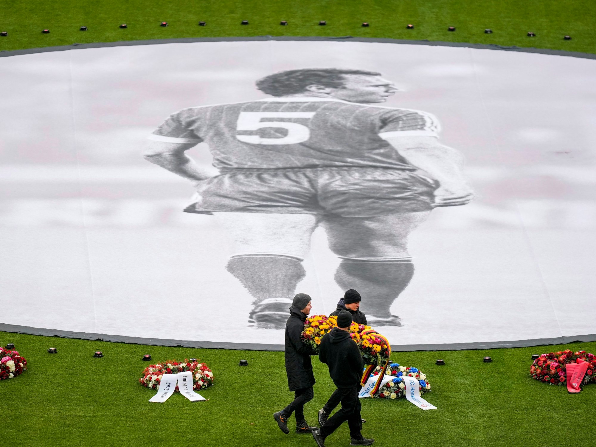 Menschen legen Blumenkränze rund um ein großes Beckenbauer-Banner im Mittelkreis der Allianz-Arena nieder.