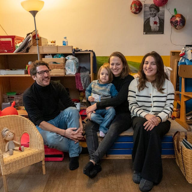 Zwei Frauen, ein Mann und ein kleines Kind sitzen in einem Wohnraum und schauen in die Kamera.