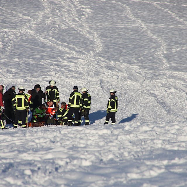 Rettungskräfte stehen auf einem tief verschneiten Hang. In ihrer Mitte knien Helfer und versorgen eine Patientin.