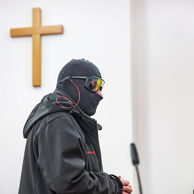 Ein Man in schwarzem Anorak, mit Sturmhaube und verspiegelter Sonnenbrille steht in einem Raum, an der Wand hängt ein Holzkreuz.&nbsp;