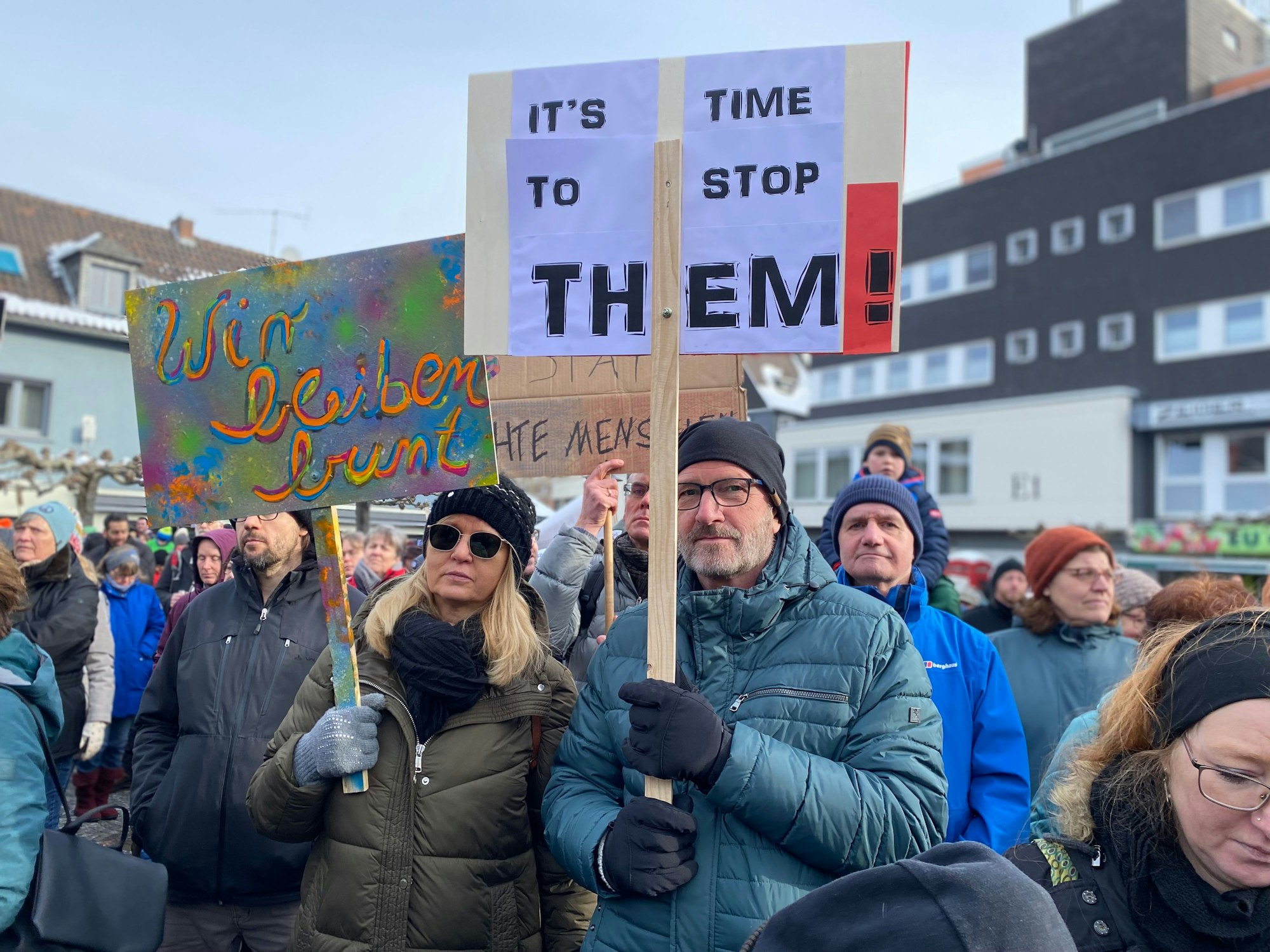 Zwei Menschen halten Plakate. Darauf steht: „Wir bleiben bunt“ und „It's time to stop them“