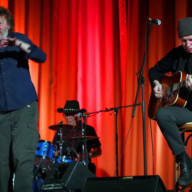 Ein Sänger, ein Schlagzeuger und ein Gitarrist auf einer Bühne vor einem roten Vorhang.