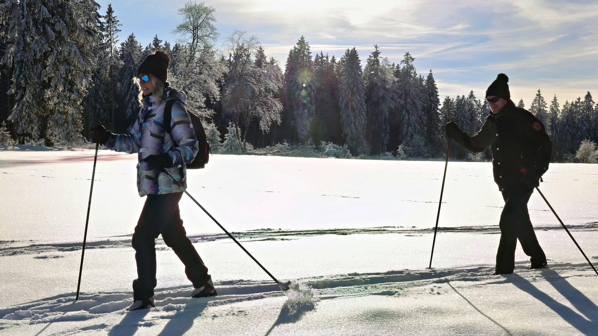 Ein junges Pärchen ist auf Skiern im glitzernden Pulverschnee unterwegs, im Hintergrund verschneite Wälder.