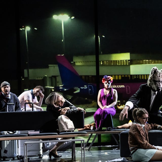 Auf der Bühne ist eine Szene in einem Flughafenterminal aufgebaut. Die Schauspieler sitzen wartend teils auch auf dem Boden. Einer trägt eine Wolfsmaske.