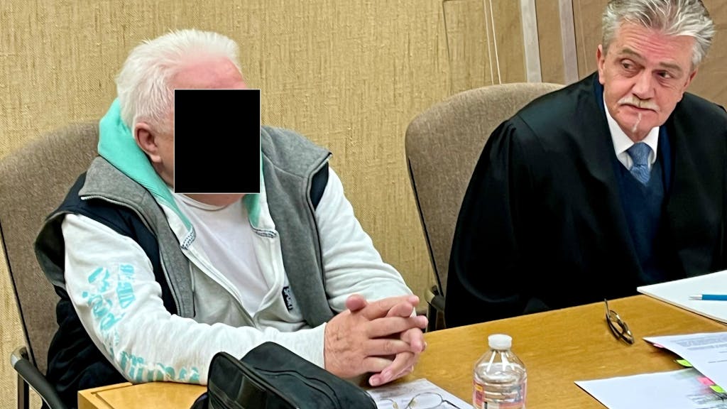 Ein Mann sitzt neben seinem Anwalt auf der Anklagebank, vor sich eine Herrenhandtasche.