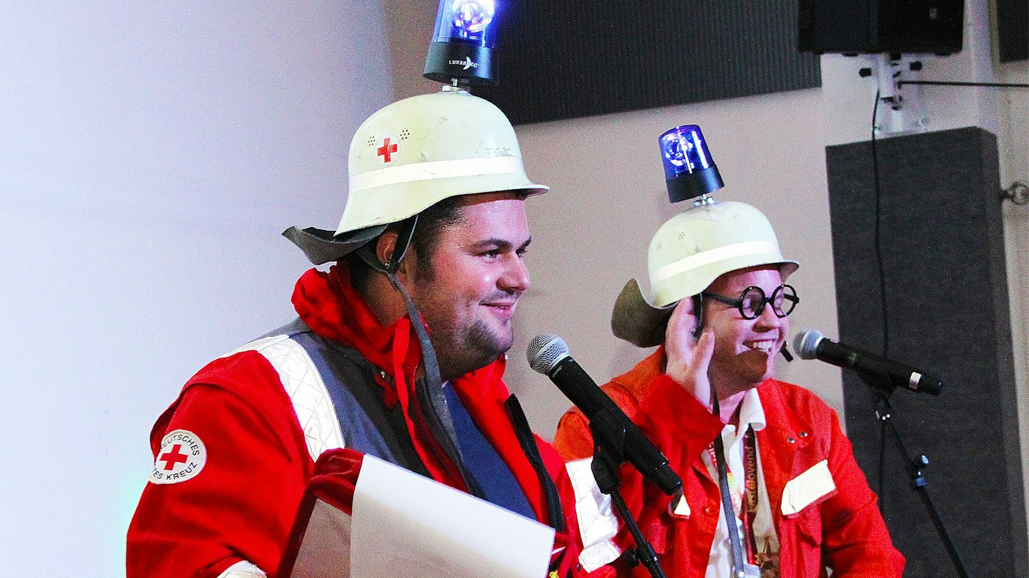 Zwei Büttenredner stehen auf der Bühne vor ihren Mikrofonen. Sie tragen Helme, die mit Blaulichtlampen versehen sind, und rote Monturen.