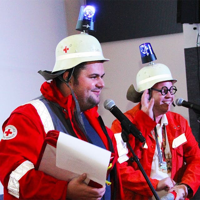 Zwei Büttenredner stehen auf der Bühne vor ihren Mikrofonen. Sie tragen Helme, die mit Blaulichtlampen versehen sind, und rote Monturen.&nbsp;
