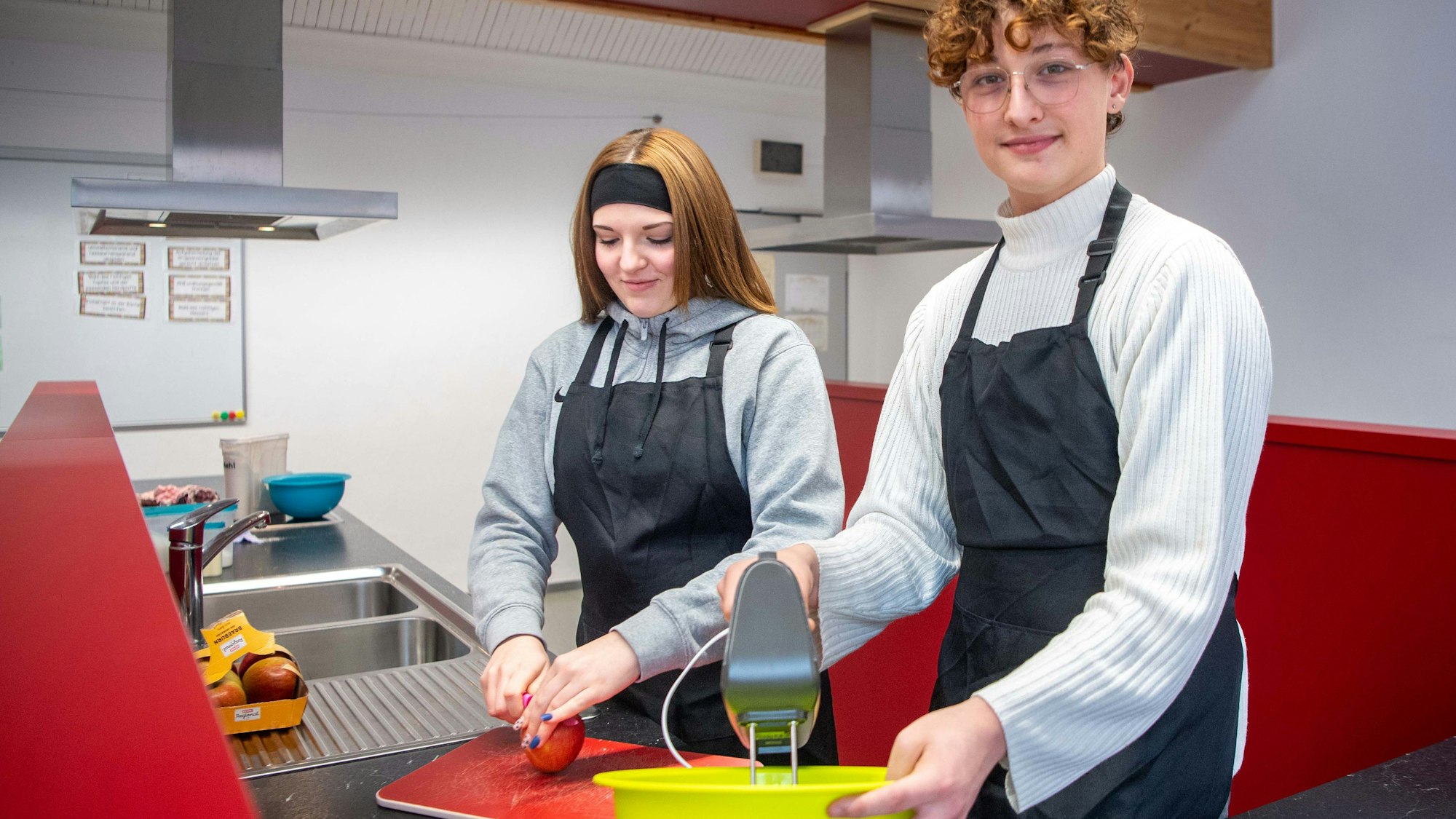 Das Bild zeigt Celina und Jakob in der Schulküche der Zülpicher Hauptschule. Celina schneidet einen Apfel, Jakob hat einen Mixer in der Hand.