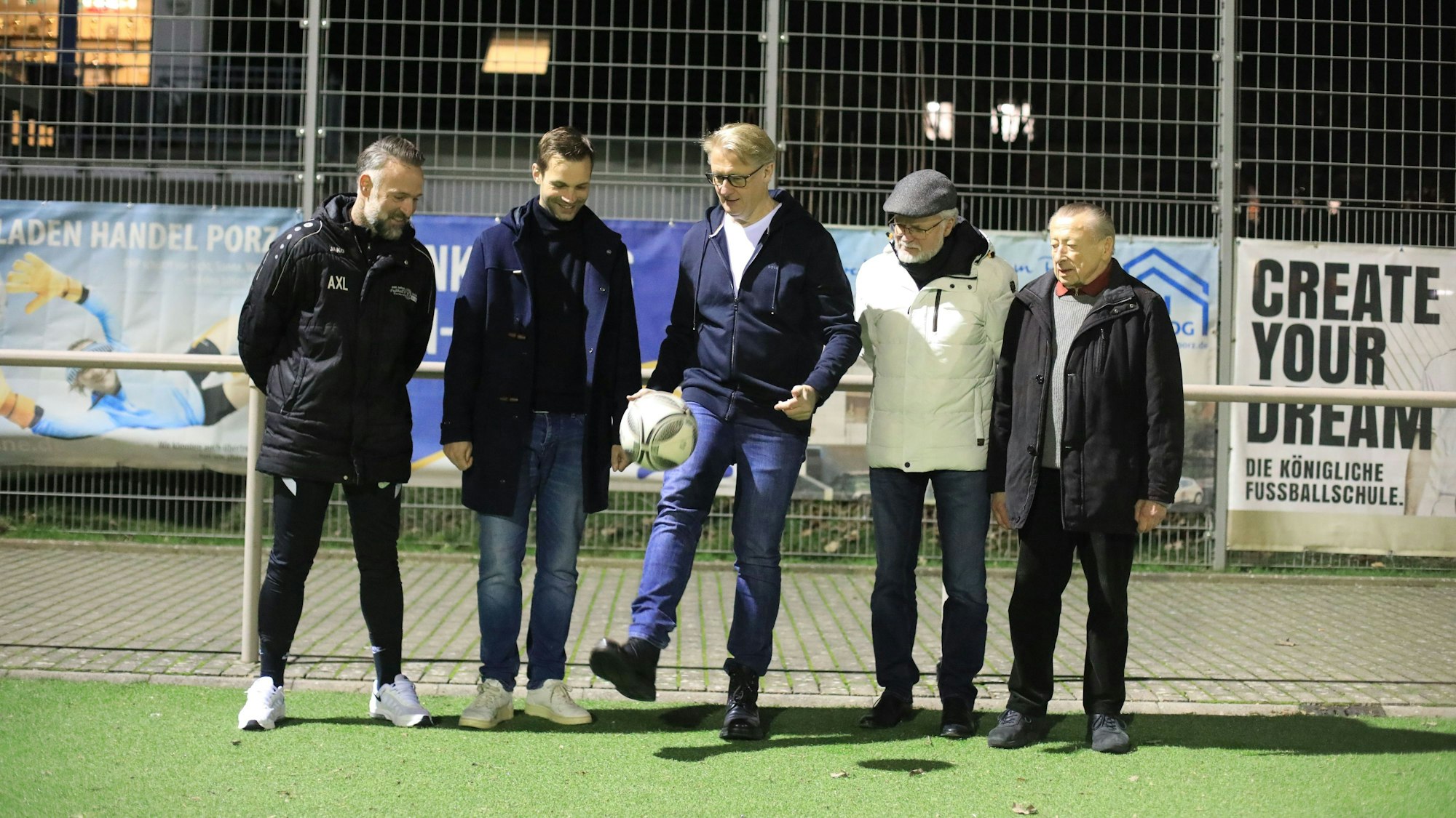 Fünf Männer stehen am Rande eines Fußballfeldes. Der Mann in der Mitte hält einen Fußball mit dem Fuß in der Luft.