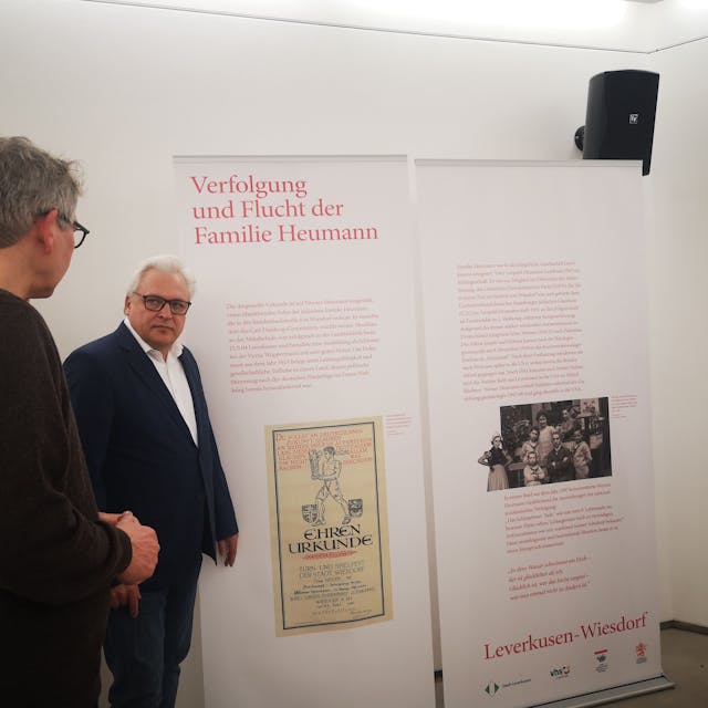 Mitarbeitende der VHS Leverkusen und der Stadt stehen vor einem Ausstellungsbanner mit dem Thema Verfolgung und Flucht der Familie Heumann
