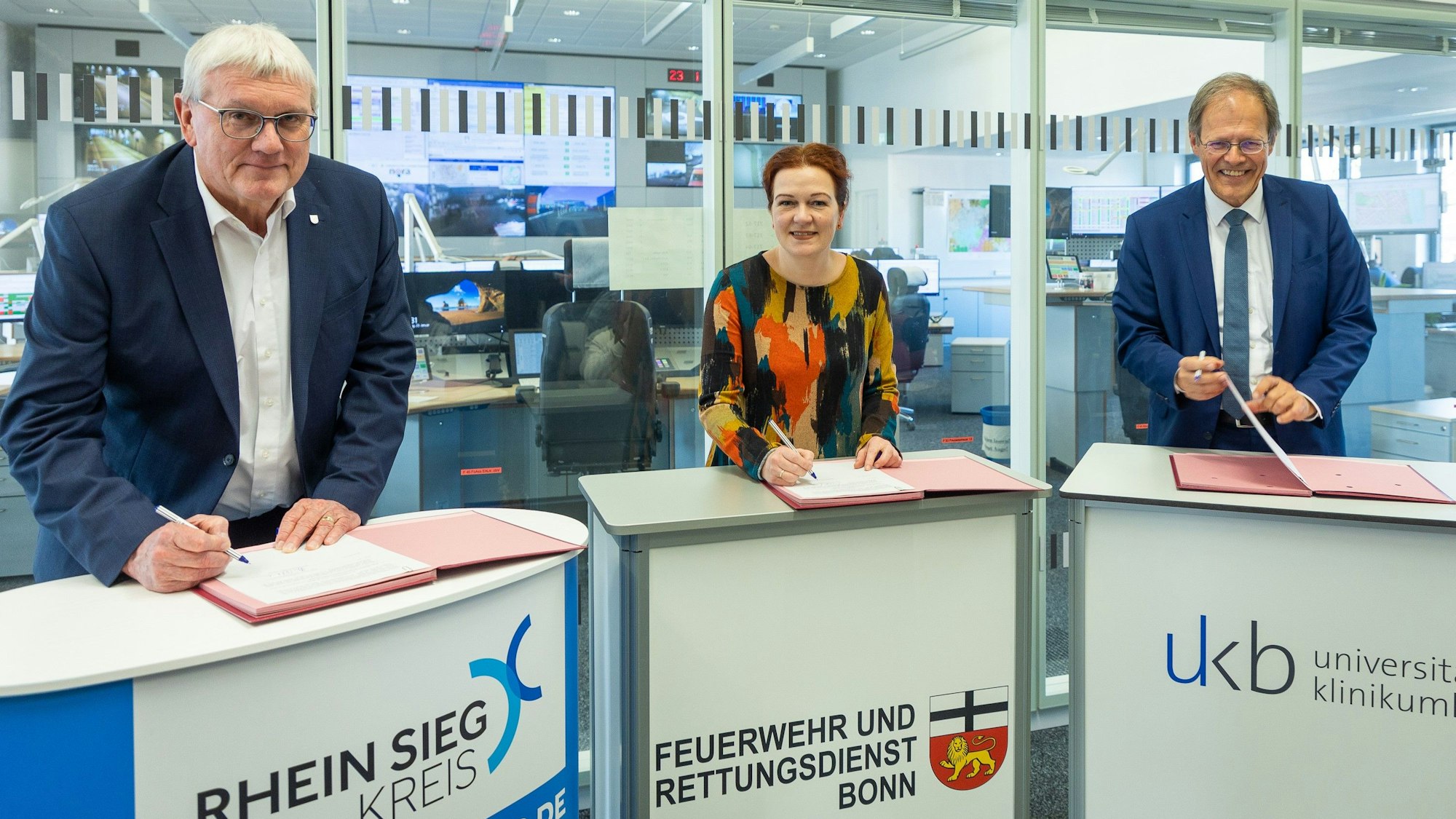 Landrat Sebastian Schuster, Bonns Oberbürgermeisterin Katja Dörner und Prof. Wolfgang Holzgreve, Ärztlicher Direktor des Universitätsklinikums Bonn (UKB), stehen in der Bonner Feuerwache 1 an Pulten und unterschreiben eine gemeinsame Vereinbarung.
