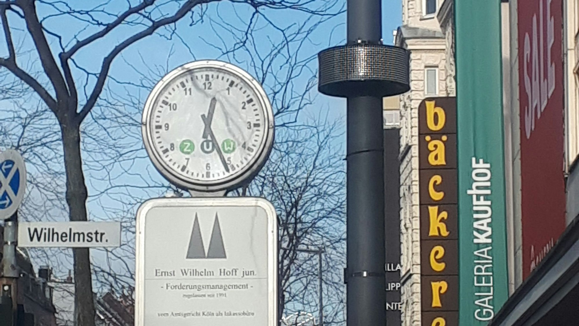 An einem Laternenmast ist ein rundes Display angebracht, daneben ist eine Uhr, ein Straßenschild und das Gebäude von Galeria Kaufhof zu sehen.