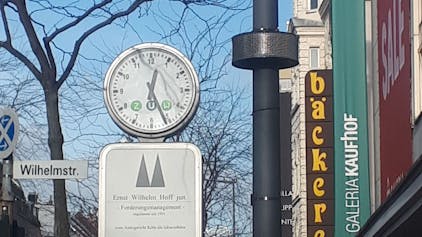 An einem Laternenmast ist ein rundes Display angebracht, daneben ist eine Uhr, ein Straßenschild und das Gebäude von Galeria Kaufhof zu sehen.