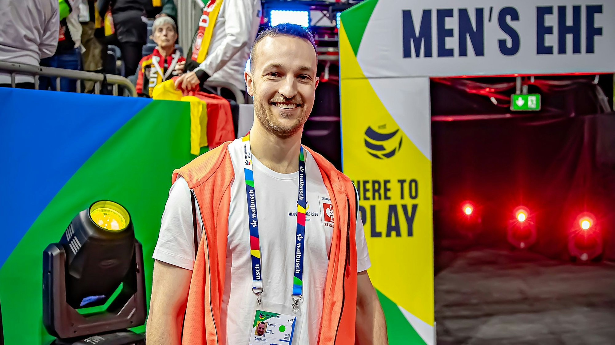 Ein junger Mann mit oranger Weste in einer Sporthalle. Im Hintergrund ist der der Schriftzug "Men's EHF" für die Handball-Europameisterschaft zu lesen.