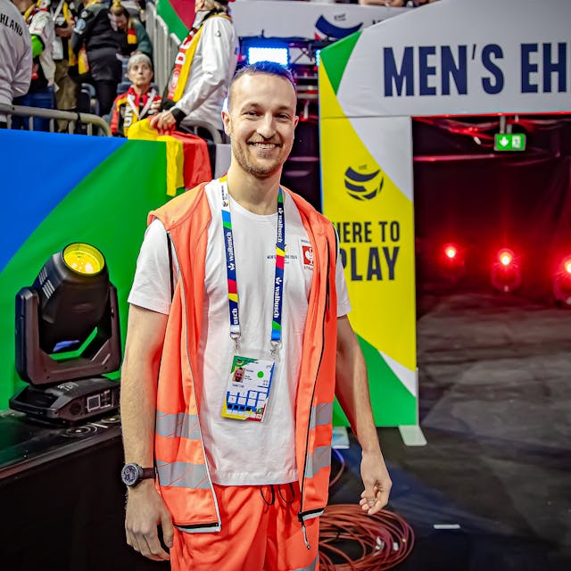 Ein junger Mann mit oranger Weste in einer Sporthalle. Im Hintergrund ist der der Schriftzug "Men's EHF" für die Handball-Europameisterschaft zu lesen.&nbsp;