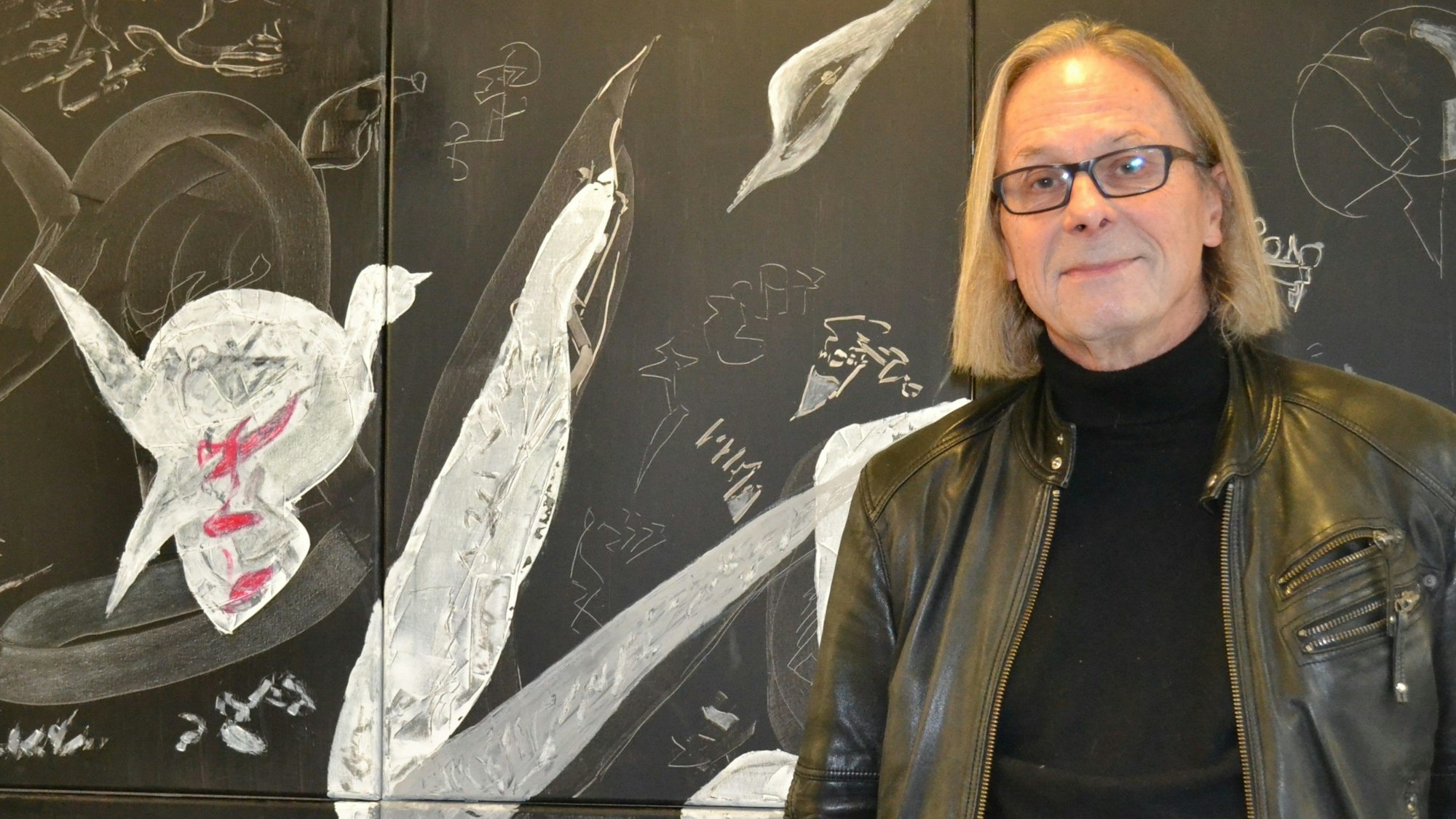 Der Künstler Hans-Heinrich Dittrich (68) mit schwarzem Pullover und Lederjacke steht vor einem seiner Bilder.