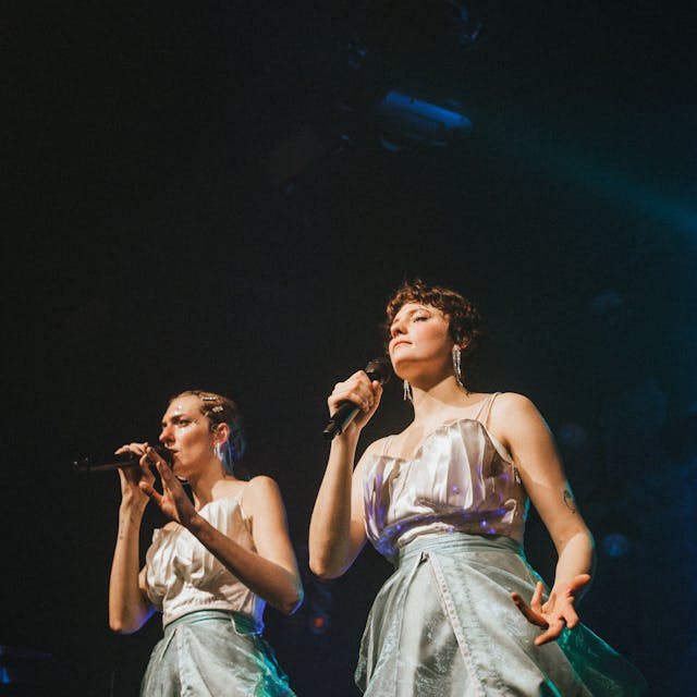 Zwei Sängerinnen in weißen Kleidern mit Glitzer im Gesicht.