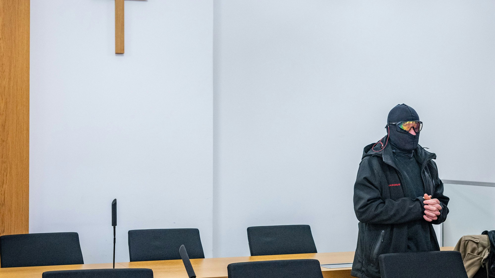 Ein Mann in schwarzer Jacke, Sturmhaube und verspiegelter Sonnenbrille steht mit gefalteten Händen in einem Gerichtssaal, an der Wand hängt ein Kreuz.