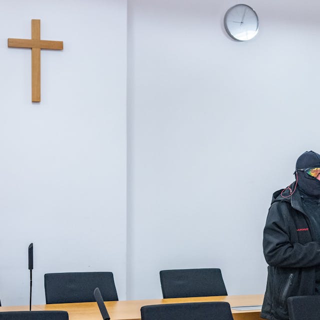 Ein Mann in schwarzer Jacke, Sturmhaube und verspiegelter Sonnenbrille steht mit gefalteten Händen in einem Gerichtssaal, an der Wand hängt ein Kreuz.