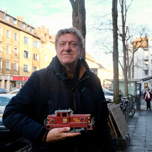 Ein Mann steht auf einem Bürgersteig an einer belebten Straße. Er hält eine Modell-Lok in der Hand.
