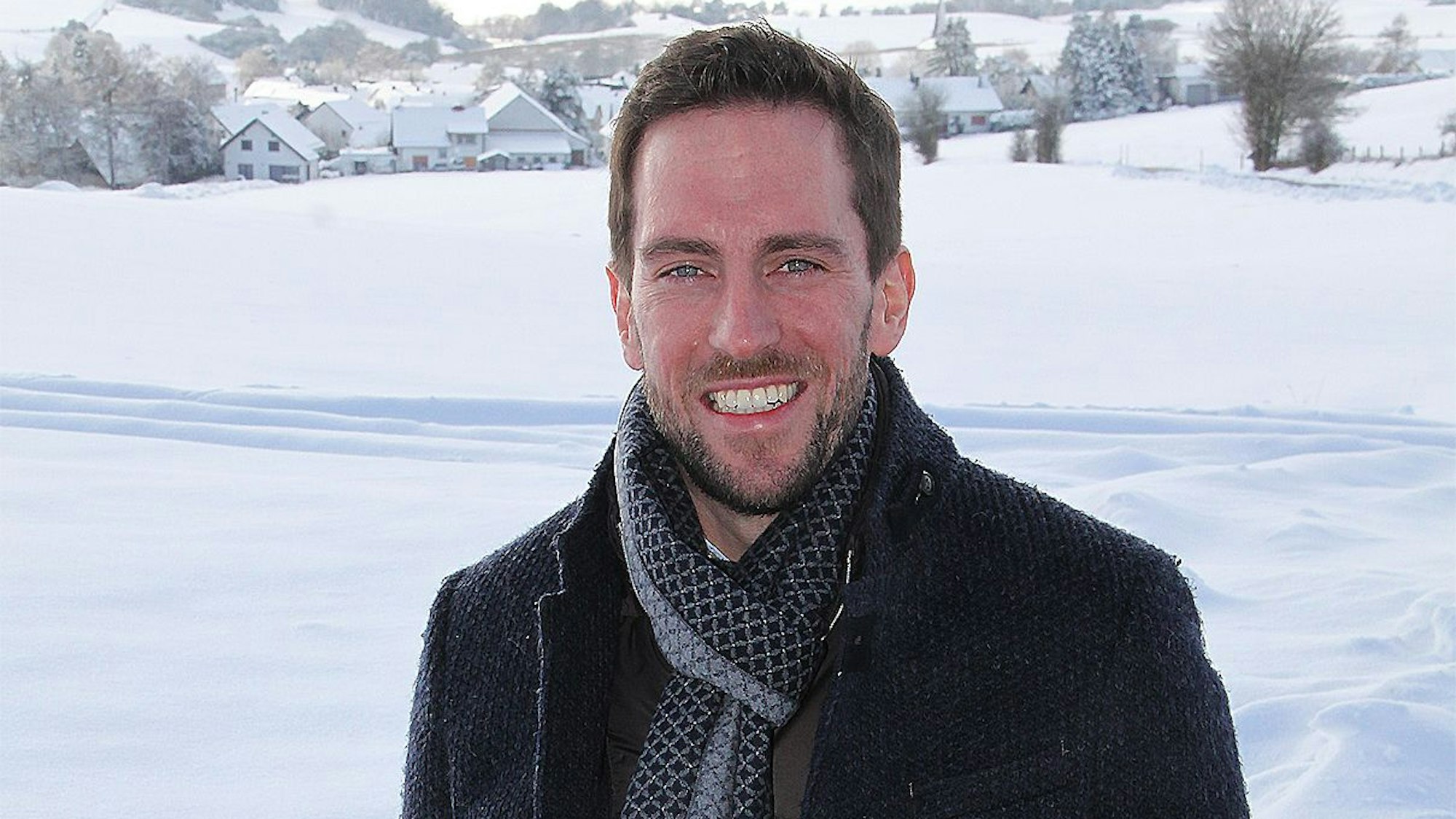 David Dreimüller in tief verschneiter Eifellandschaft. Er trägt einen Schal und einen Wintermantel.