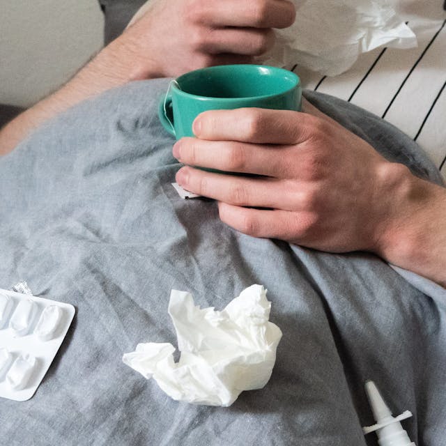 Ein Mann liegt mit Taschentüchern, Teetasse, Nasenspray und Tabletten im Bett (gestellte Szene).