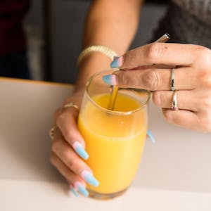 Eine Frau mit bunten Fingernägeln trinkt mit einem Strohhalm aus einem Glas Orangensaft.&nbsp;